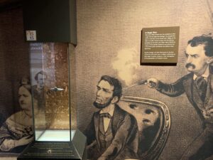 A museum display, including a small pistol in a case, a blown-up image of that pistol being used to assassinate President Abraham Lincoln, and a sign about the weapon. The sign stages, "A Single Shot After John Wilkes Booth shot the president at close range with his single shot deringer, he dropped the pistol on the floor of the theatre box. Pulling out his knife, he swiftly moved to jump over the balcony, grappling momentarily with Major Henry Rathbone, a guest of the Lincolns. Rathbone was severely cut in the brief scuffle with Booth and suffered major blood loss. Booth's deringer was later discovered on the floor of the box and turned over to military authorities as they began to gather evidence in their investigation of the assassination of President Lincoln."