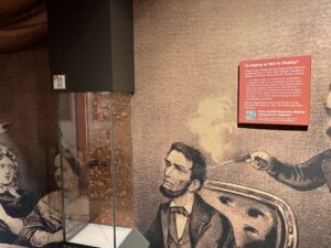 A museum display, including a small pistol in a case, a blown-up image of that pistol being used to assassinate President Abraham Lincoln, and a revised sign about the weapon. The sign stages, "A Single Shot To Display or Not to Display? After Booth killed Lincoln, the War Department kept the murder weapon. In 1931, they refused to display it and other items at Ford's Theatre, stating: '...the relics should not be allowed to be displayed to the public under any circumstances, on the theory that they would create interest in the criminal aspects of the great tragedy, rather than in the historical features thereof, and would have more of an appeal for the morbid or weak-minded than for the students of history. ... the Lincoln relics should not be placed upon exhibition anywhere.' The War Department transferred the gun to National Park Service in 1940. It has been displayed here since. How should museums display weapons of political violence? Tell us what you think! Scan this code or go to: www.surveymonkey.com/r/WeaponDisplay"