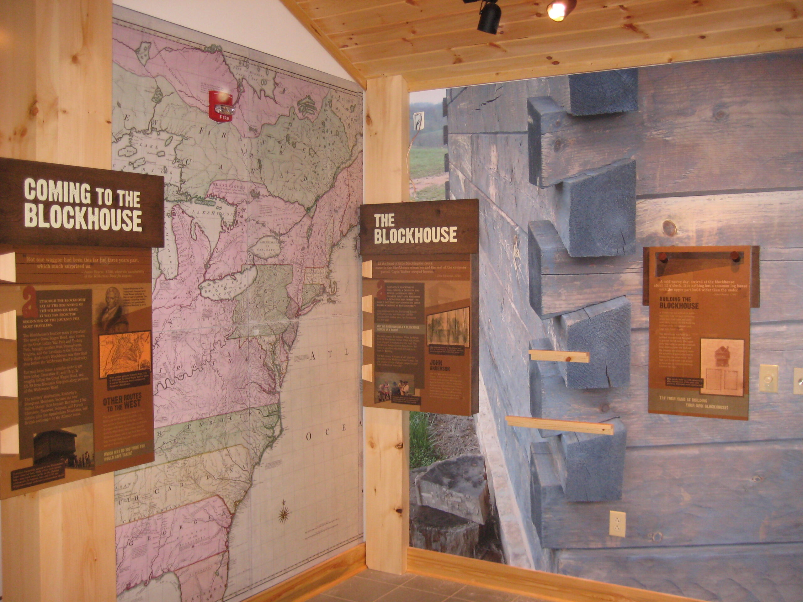 Two museum exhibition panels in a corner. One says simply "The Blockhouse," while the other says, "Coming to the Blockhouse." Background panels are a map of eastern North America circa 1775 and a photograph of the corner of a log cabin.