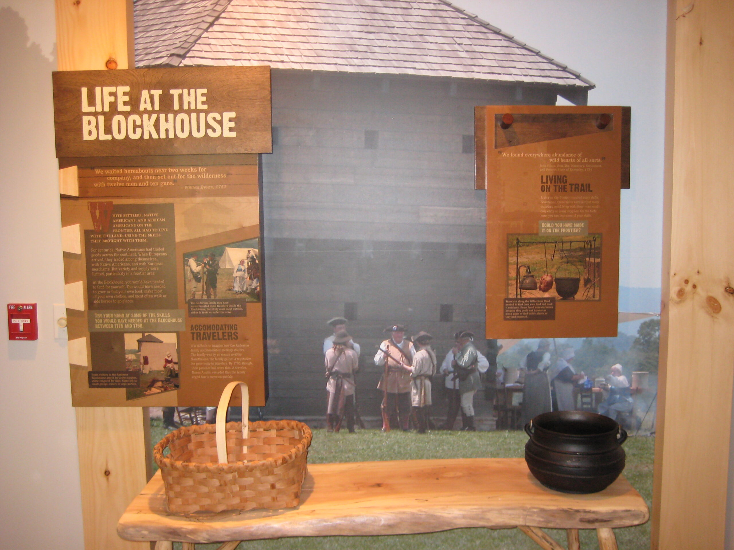 An exhibition graphic titled "Life at the Blockhouse," in front of a picture of 1700s militiamen in front of a log of a blockhouse. A basket and an iron pot is displayed at front.