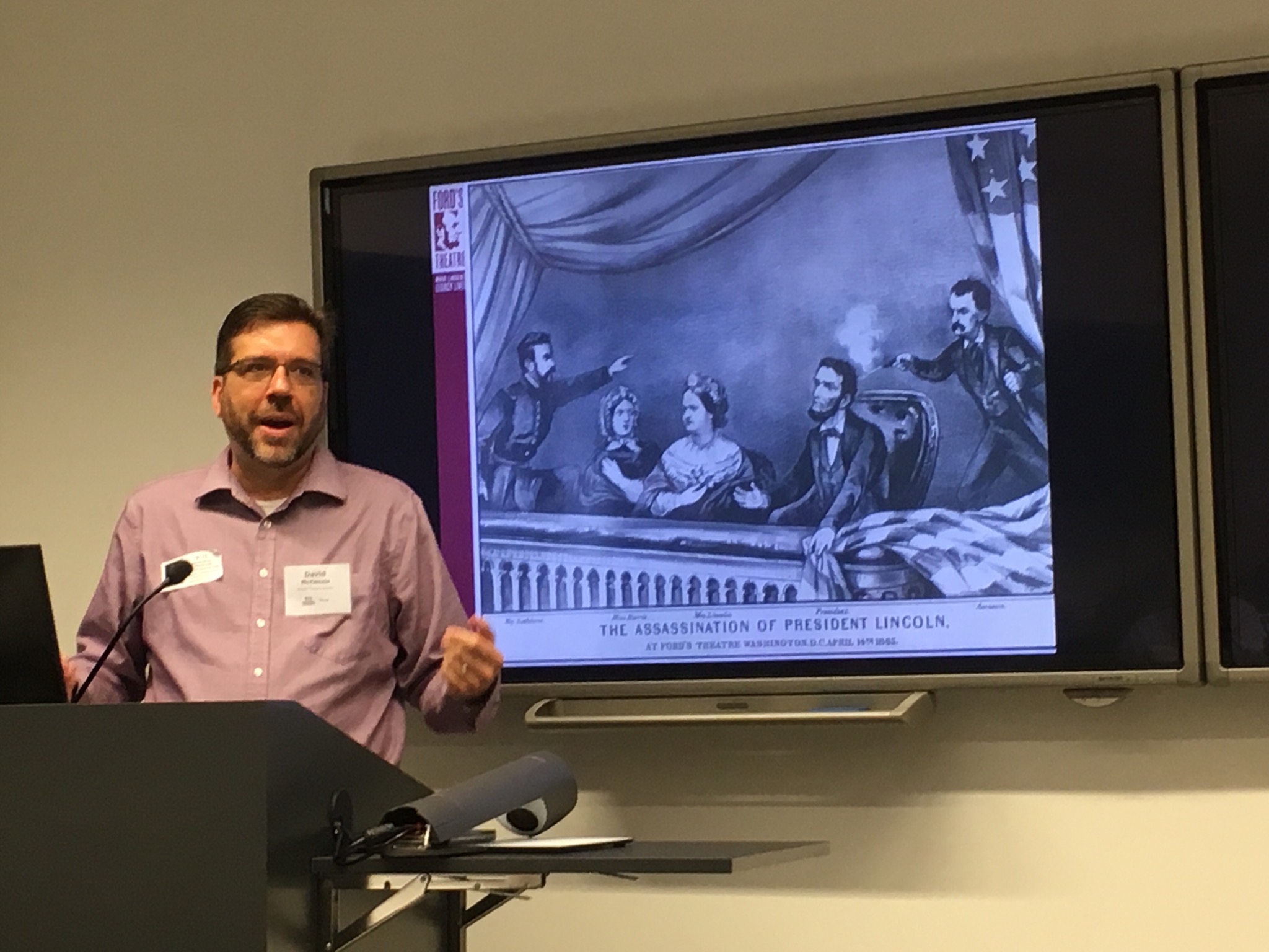 A man speaks in front of a picture of the assassination of President Abraham Lincoln.